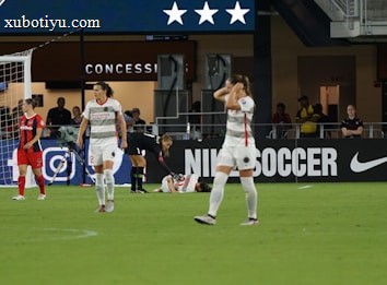 A soccer match scene shows players on a field, some in red and others in white uniforms. One player is injured and lying on the ground while another kneels beside them. The area behind features a concession stand with a large advertisement for Nike Soccer. Spectators and staff are visible in the background.