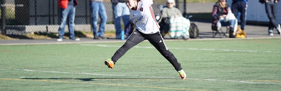 A person is mid-air on a sports field, appearing to be involved in an athletic activity. Several spectators are on the sidelines, some seated and others standing, with one individual capturing the event with a camera. The background includes a fence and various casual observers, some of whom are wearing winter clothing.