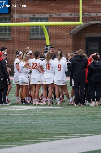 A group of athletes wearing sports uniforms huddles on a grassy field, likely preparing or strategizing for a game. The players are surrounded by additional team members or coaches wearing black jackets. A goalpost and net are visible in the background, along with a brick building.