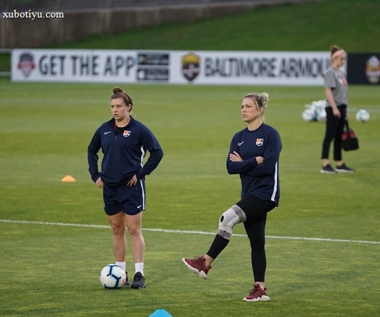 Two individuals are standing on a grassy sports field, each wearing dark athletic clothing with a logo. One person is balancing on one leg with a brace on their knee, while the other stands with hands on hips. Soccer balls and markers are visible in the background, along with a person collecting equipment.